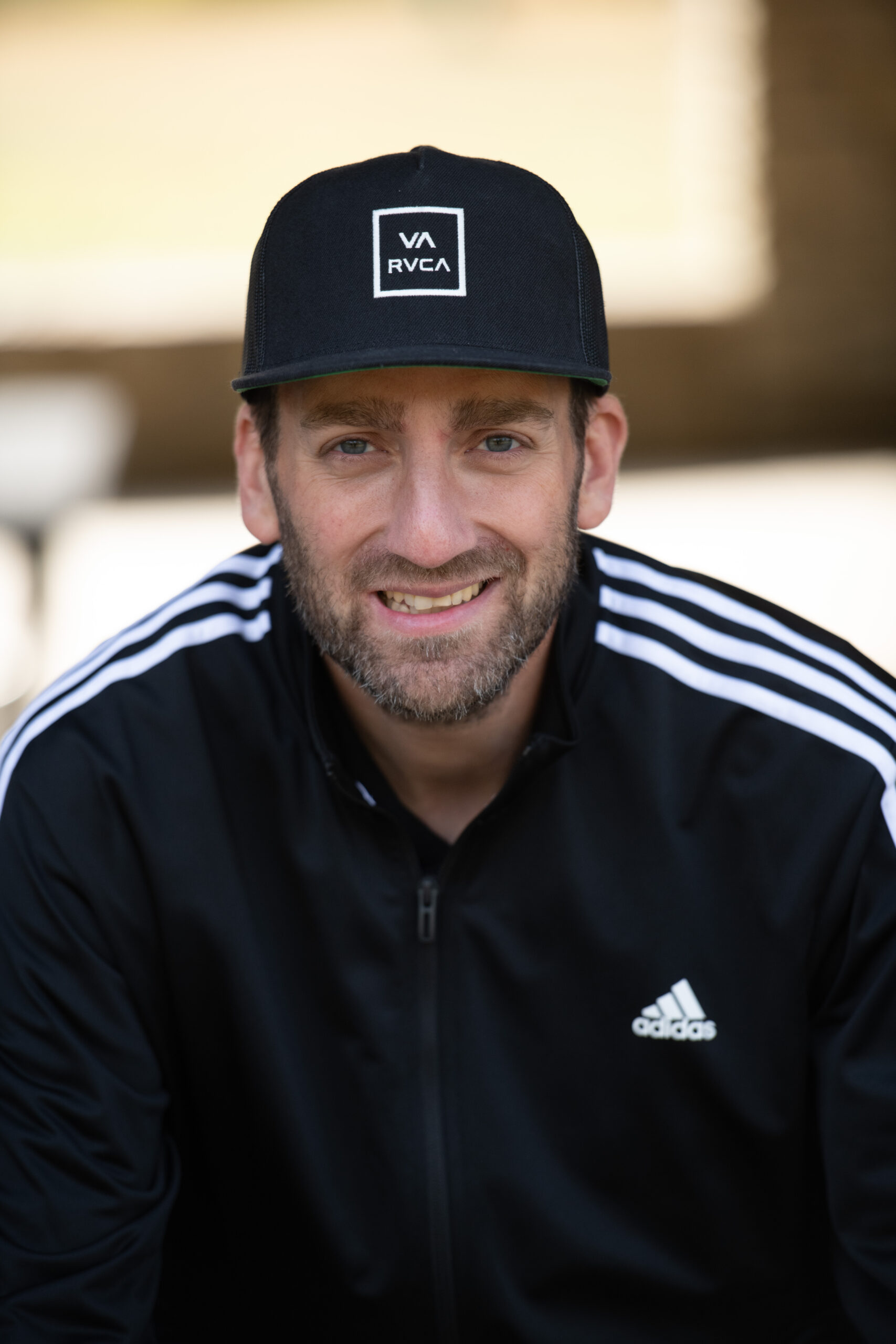 Jamie Edwards smiling with a black baseball cap and a black Adidas track suit sits in front of a blurred out harth